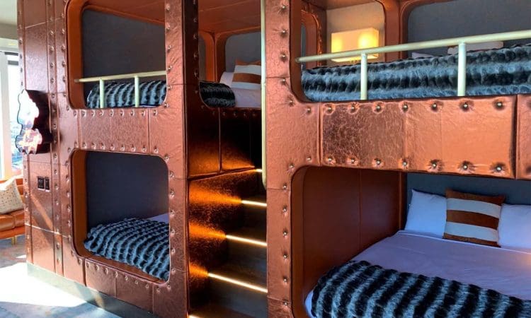 Four built-in copper-colored bunk beds with striped pillows and faux fur throws, separated by a central staircase with illuminated steps.