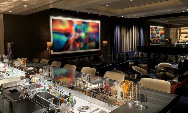 Modern lounge bar with a sleek countertop, various bottles and glassware, plush seating, and large abstract artwork on the walls. The lighting is warm and ambient.