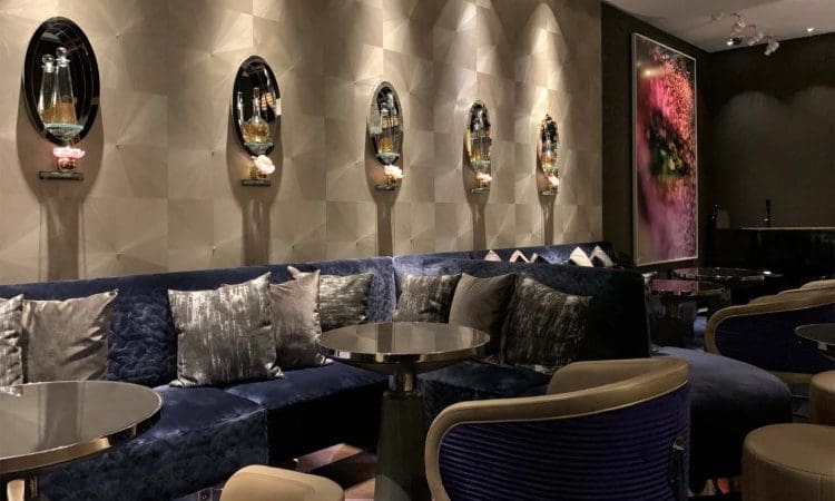 Modern lounge with blue velvet seating, round tables, decorative wall mirrors holding bottles, plush cushions, and dim lighting. Large abstract art hangs on the wall.