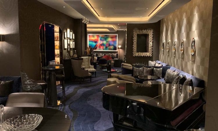 Modern lounge with dim lighting, a grand piano, plush seating, artwork on the walls, and a bar area in the background.