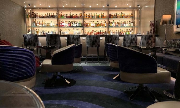 Modern bar interior with a backlit wall of assorted bottles, high chairs at the bar, and rounded lounge chairs in the foreground on a patterned carpet.