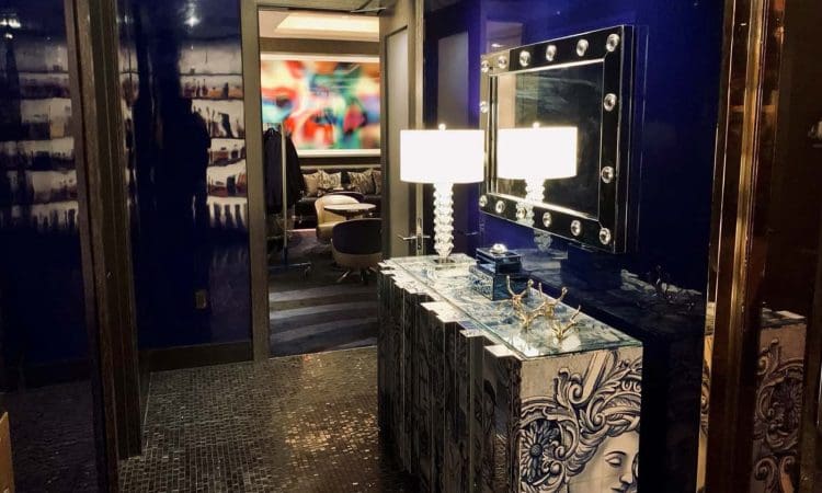 A modern hallway with glossy blue walls, a decorative console with artistic face designs, two lit lamps, a mirror, and an open door revealing a colorful room.