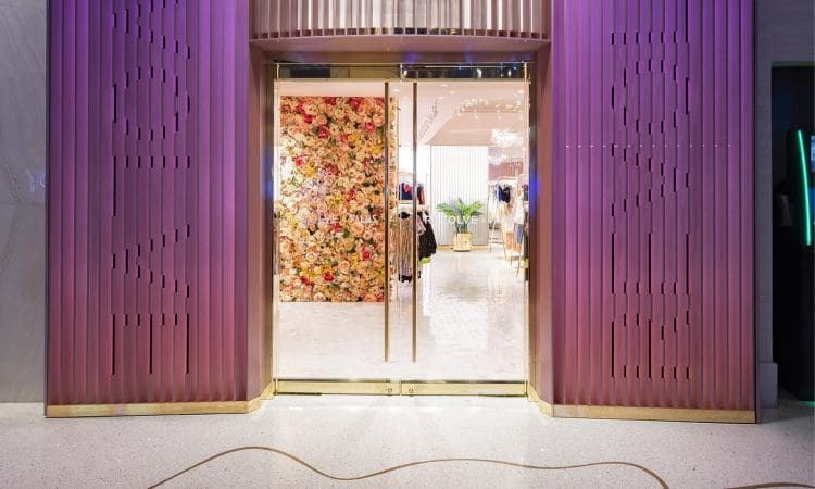 Glass double doors with gold trim open to a boutique interior featuring a wall covered in colorful artificial flowers, mannequins, and clothing displays.