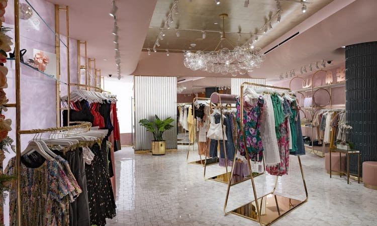 A modern boutique with pink walls and gold clothing racks, displaying dresses and garments on hangers, with decorative lighting and plants.