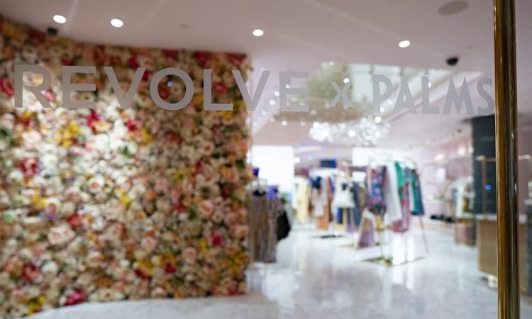 Glass door with "REVOLVE x PALMS" text, showing a fashion boutique interior with floral wall and racks of clothing in the background.