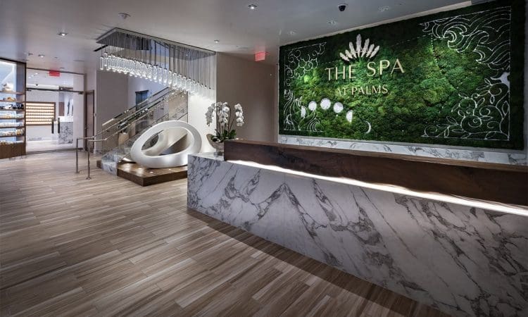 Reception area of The Spa at Palms with a marble desk, green wall sign, modern decor, wooden flooring, and stairs in the background.
