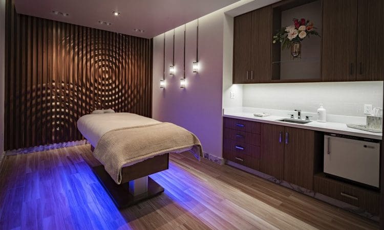 A modern spa room with a massage table, soft lighting, wood paneling, blue accent lighting on the floor, and built-in cabinets with a small sink and flower arrangement.