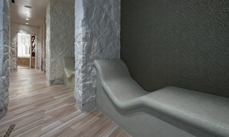 A hallway with light wood-look tile flooring, textured white stone walls, and a curved tiled lounge chair against a mosaic-tiled wall.
