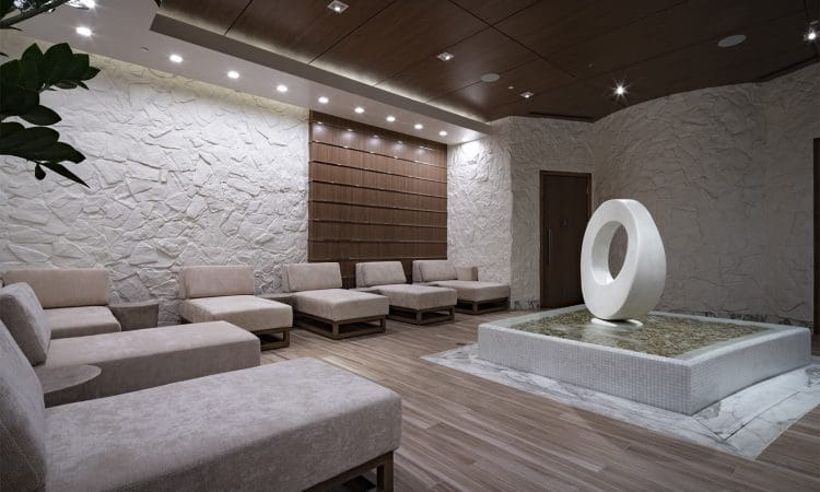 Modern waiting room with beige lounge chairs, textured stone walls, wood accents, and a circular abstract sculpture on a tiled platform in the center.