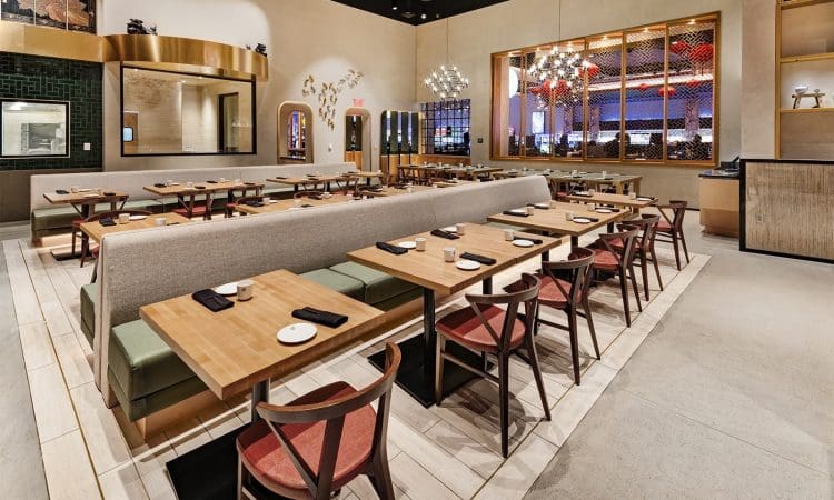 Modern restaurant interior with wooden tables, red and green chairs, beige walls, decorative lighting, and neatly arranged place settings. The space appears clean and organized.