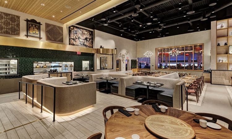 Modern, spacious restaurant interior with wooden and beige furnishings, round and rectangular tables, decorative wall art, and ambient lighting. No people are present.