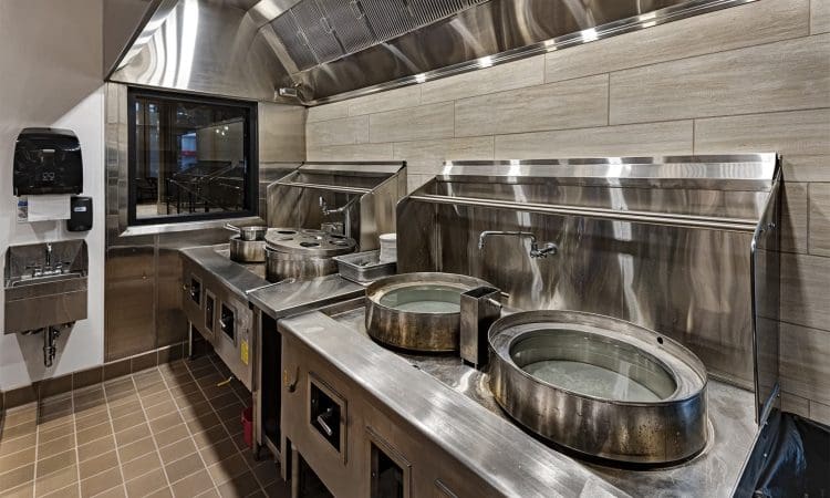 A commercial kitchen with three large wok stations, stainless steel surfaces, and a wall-mounted sink on the left.