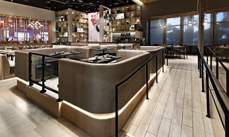Modern restaurant interior with beige booth seating, wooden flooring, decorative shelving, and large windows letting in natural light. Tables are set and lighting fixtures hang from the ceiling.