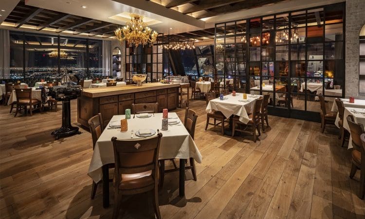 Elegant restaurant interior with wooden floors, set tables, chandeliers, and large windows offering a city view at night.