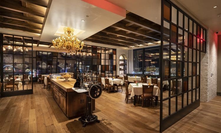 Elegant restaurant interior with wooden floors, chandeliers, large windows, and neatly set tables, featuring a central serving station and city lights visible outside.
