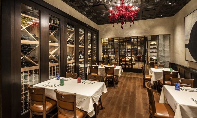 Elegant restaurant dining room with wine racks, wooden floors, tables with white tablecloths, leather chairs, and a red chandelier, with a glass wall showing another room in the background.