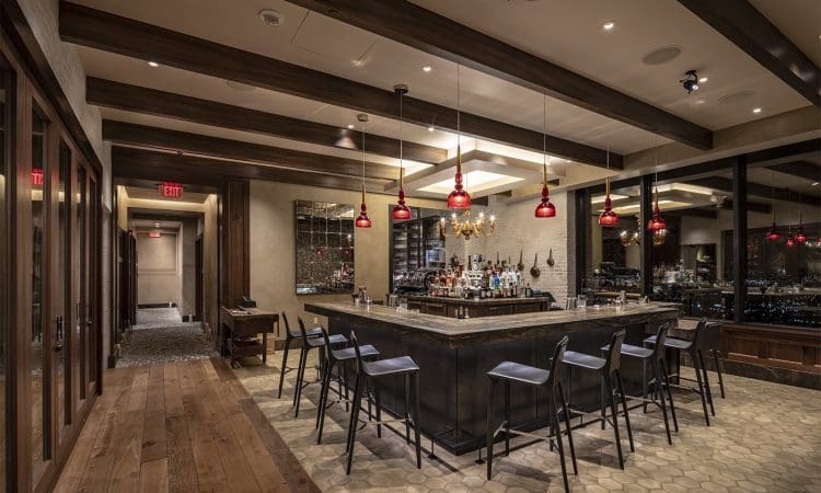 Modern bar with high-top chairs, pendant lights, and large windows overlooking a city at night. Wooden floors and ceiling beams create a warm, upscale atmosphere.