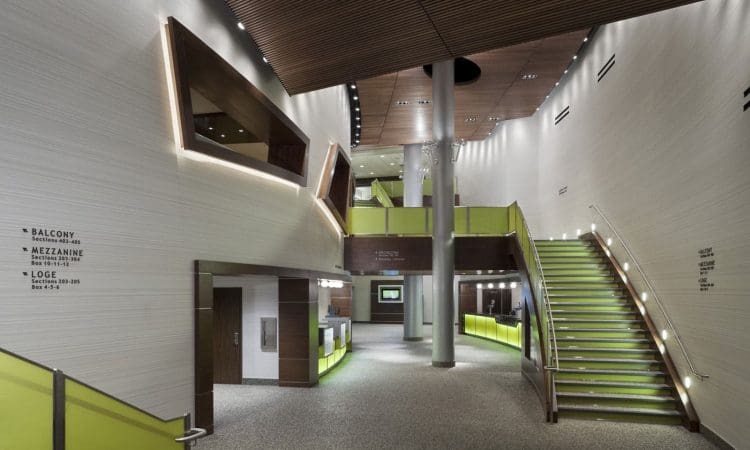 Modern theater lobby with lime green accents, wide staircase, mezzanine level, wood ceiling panels, and signage directing to balcony, mezzanine, and loge areas.