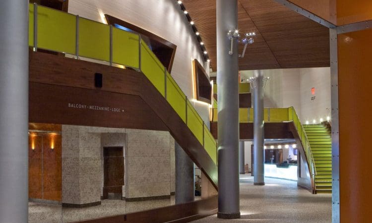 Modern interior with green-accented staircase and balcony, exposed support columns, textured walls, and a lit reception area in the background.