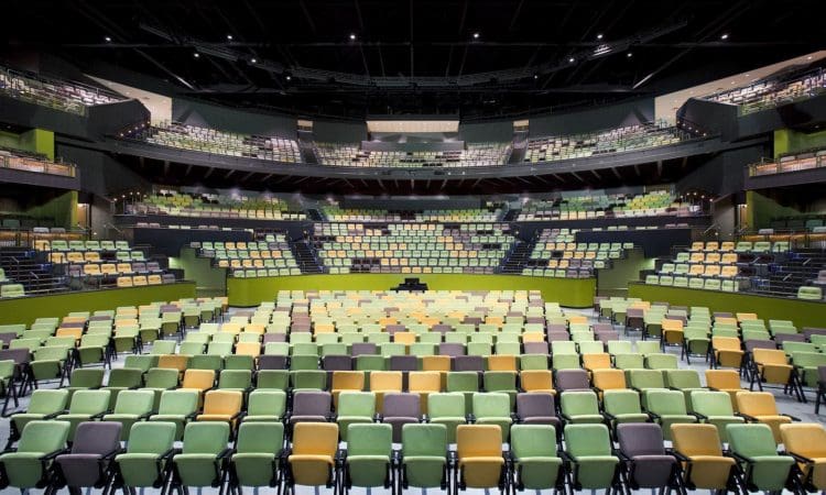 A large, modern auditorium with multiple tiers of seating in green, yellow, and tan tones, viewed from the stage facing the empty seats.