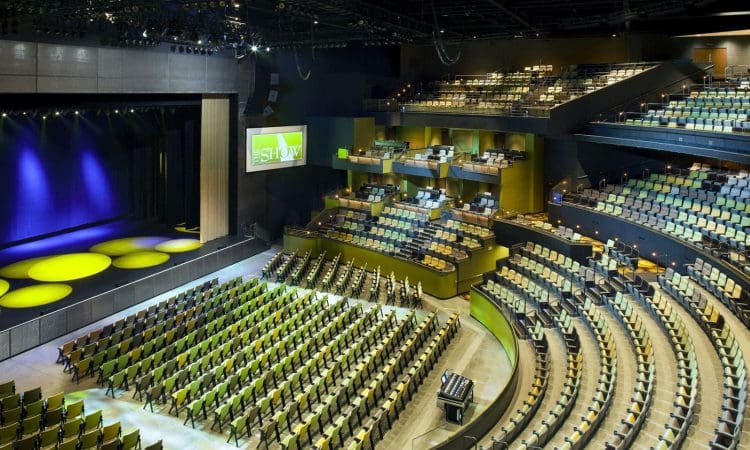 A large, modern theater auditorium with rows of empty seats, a stage lit with yellow spotlights, and a screen displaying the word "SHOW.