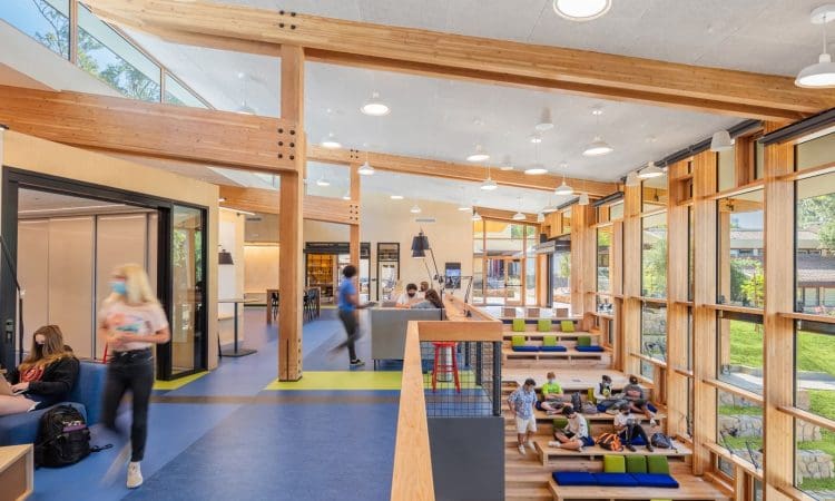 Modern indoor common area with wood beams, large windows, and blue flooring. People are walking, sitting, and socializing in various seating areas. Natural light fills the space.