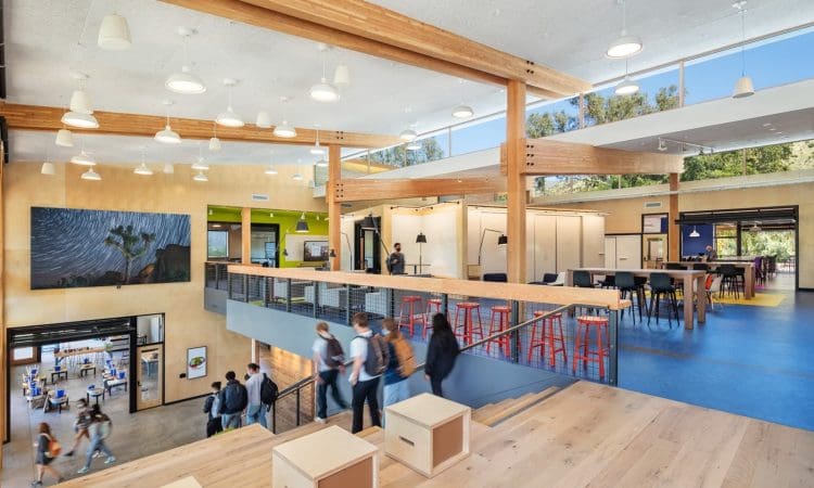 Open, modern interior of a building with wood beams, seating areas, people walking, large windows, and artwork on the wall.