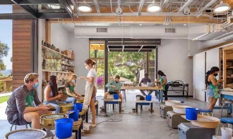 People working at pottery wheels in a bright, modern studio with large windows and shelves displaying ceramic pieces. Some participants wear masks and aprons.