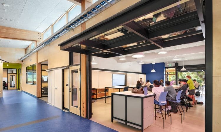 A group of people sit around tables in a modern classroom, listening to a presenter next to a screen, with large windows and an open garage-style door.