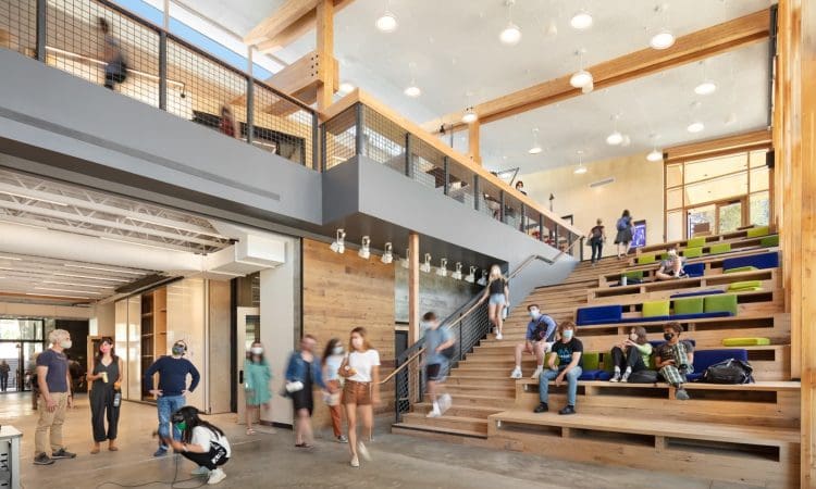 Students and adults walk and gather in a modern, open atrium with large wooden steps, seating, and an upper level with a railing. Natural light fills the space.