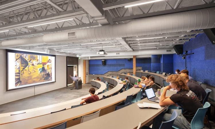 Students sit in a modern lecture hall with tiered seating, watching a presentation projected onto a large screen at the front of the room.