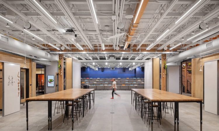 A modern open workspace with four wooden tables and stools in the foreground, and tiered seating and exposed ceiling pipes in the background. A person walks through the center.