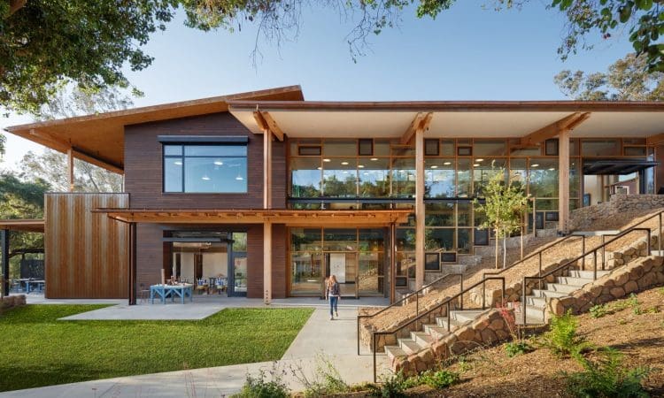 A modern two-story building with large windows, wooden accents, and a sloped roof, surrounded by greenery and a stone staircase.