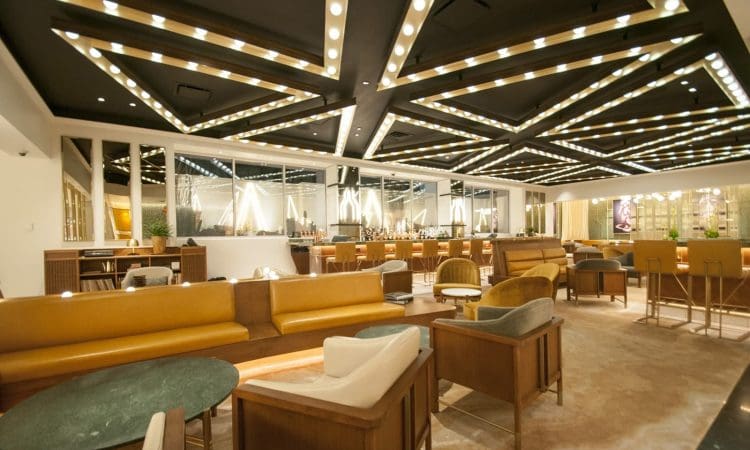 Modern lounge with geometric ceiling lights, brown leather sofas, armchairs, and tables arranged in a spacious, well-lit area with a mirrored wall and decorative plants.