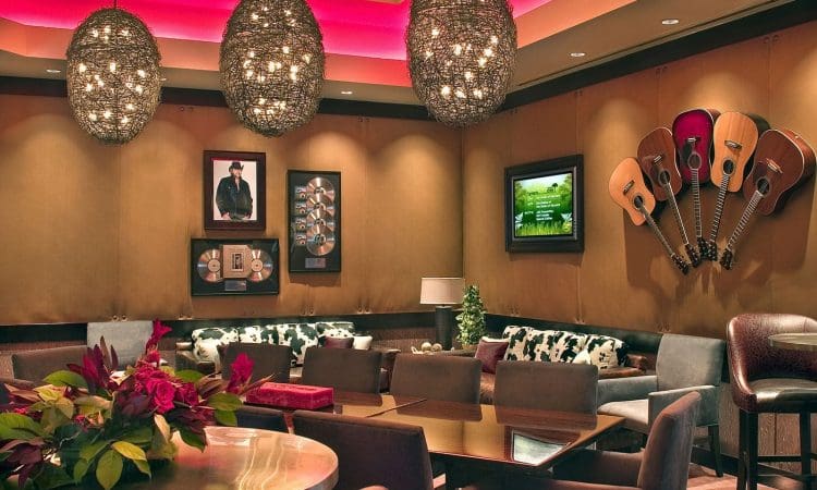 A lounge with modern lighting, wall-mounted guitars, framed music memorabilia, a TV, assorted chairs and tables, and a floral centerpiece on one table.