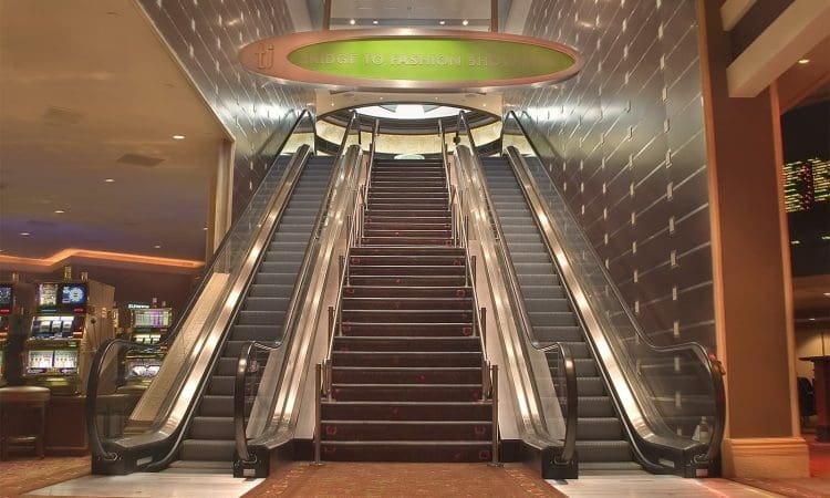 A wide staircase flanked by two escalators leads up to a sign reading "Bridge to Fashion Show Mall" inside a brightly lit building.
