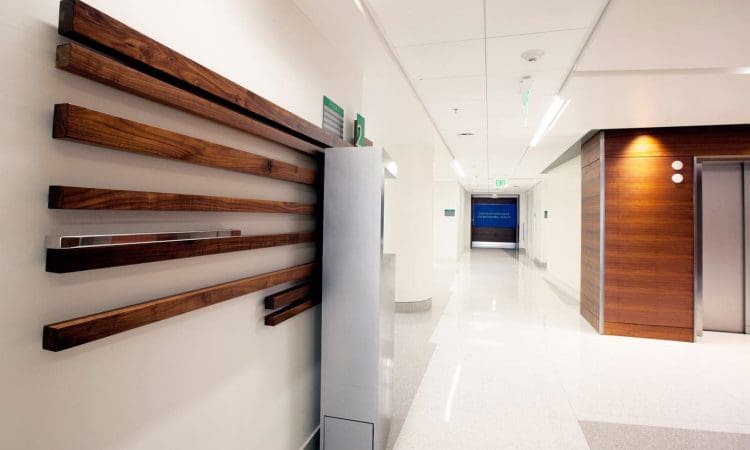 Modern hospital hallway with light walls, polished floors, wooden accents, and an elevator on the right. A decorative wood panel and sanitizer dispenser are mounted on the left wall.