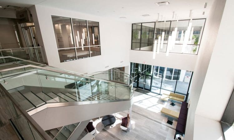 Modern office lobby with glass walls, staircase, hanging lights, lounge seating, and large windows letting in natural light.