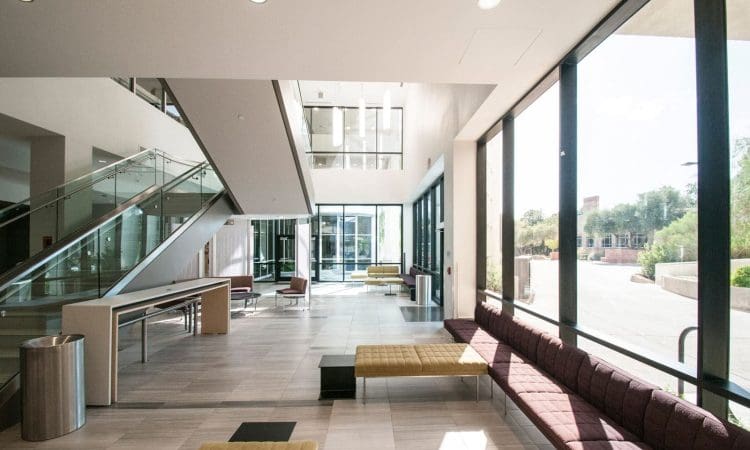 Modern building lobby with large windows, natural light, benches, tables, a trash can, and a staircase on the left.