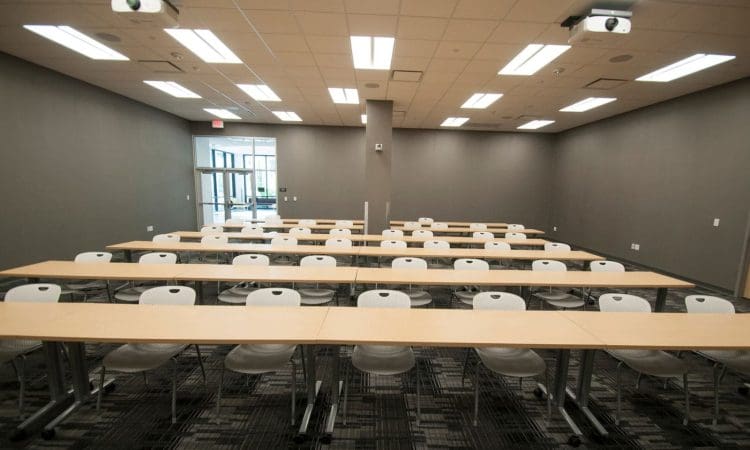 An empty classroom with rows of light-colored tables and white chairs, overhead lights, projectors on the ceiling, and a door at the back.