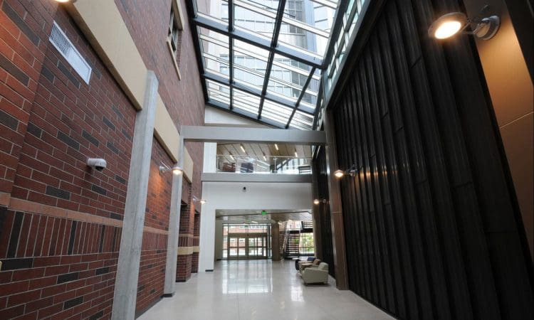 A modern building lobby with brick and dark panel walls, glass ceiling, overhead lights, and a seating area near glass entrance doors.