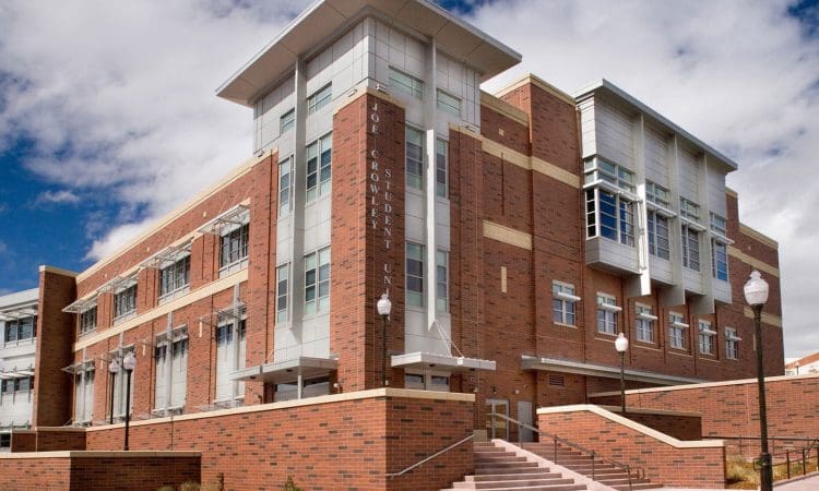 A modern brick and glass building with large windows, multiple stories, and stairs leading to the entrance, set against a partly cloudy sky.