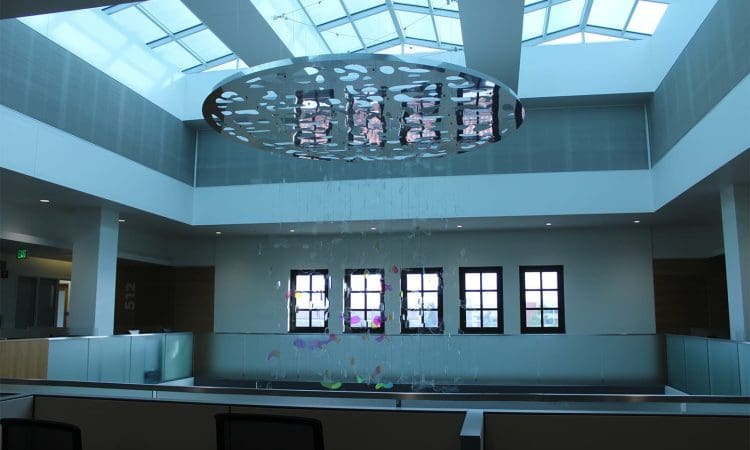 Bright atrium with a large skylight, modern hanging art installation, and multiple square windows along the back wall above office cubicles.