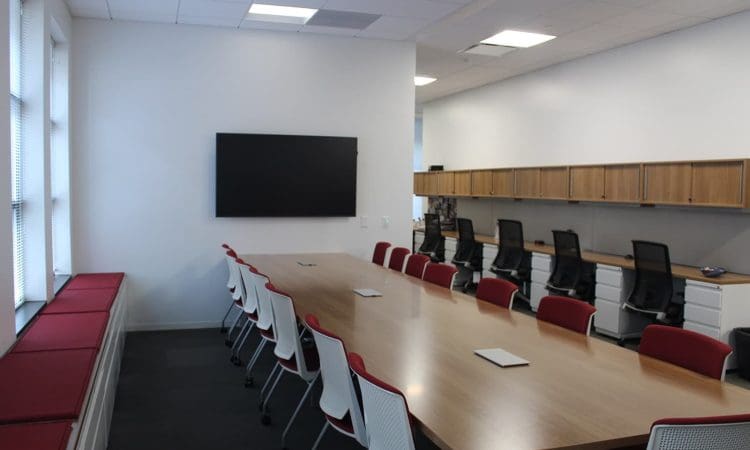 A modern conference room with a long wooden table, red and white chairs, a wall-mounted screen, and workstations with office chairs along the back wall.