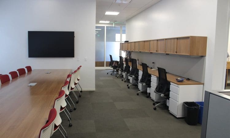 Modern office space with a long conference table and chairs on the left, individual desks with office chairs and storage on the right, and a wall-mounted screen.