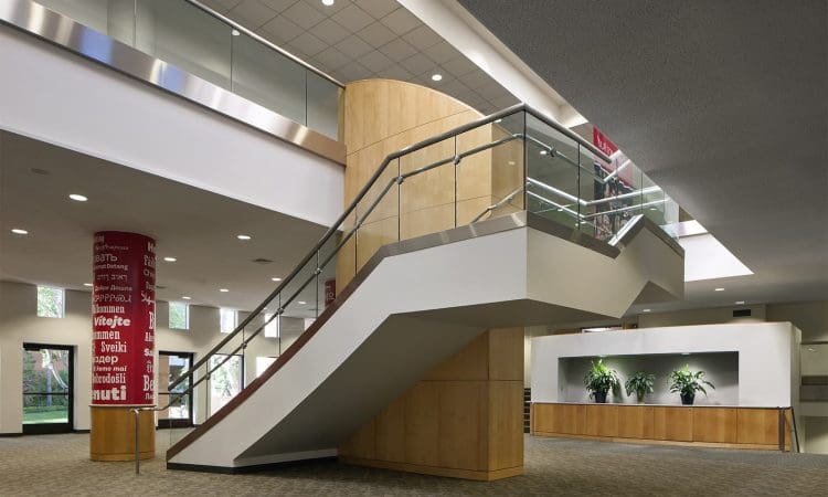 Modern indoor space with a wide staircase, glass railing, carpeting, two red columns with white text, and a reception area with potted plants in the background.