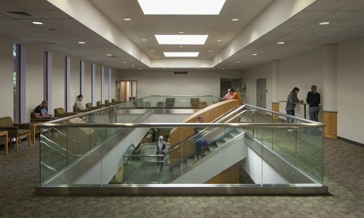 Interior view of a modern building with a central staircase and glass railings; people are seated and standing around the open upper level.