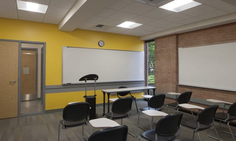 A classroom with rows of black chairs and white desks facing a whiteboard, podium, and teacher’s desk, with yellow and brick walls and a window showing greenery outside.