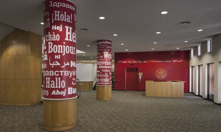 Lobby with red pillars displaying greetings in multiple languages, patterned carpet, wood paneling, a reception desk, and a red accent wall with a school emblem.