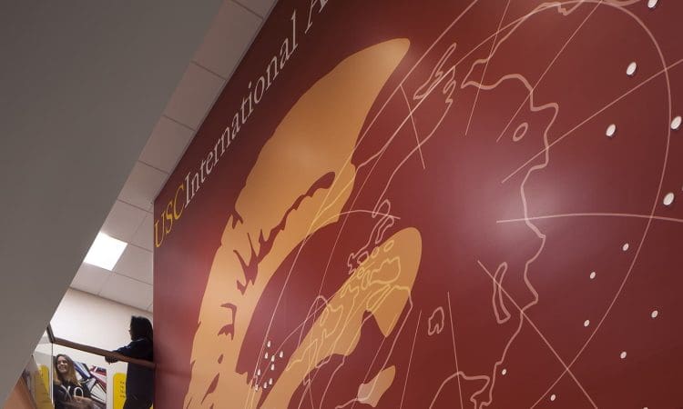 Two people stand near a staircase beside a large mural with a map and the words "USC International Academy" on the wall.